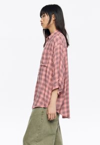Camisa a cuadros de color rosa claro con botones y cuello, con mangas enrolladas y un ajuste holgado. Tejido suave con un bolsillo en el lado izquierdo.