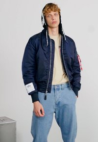 Mornarska bombarder jakna s patentnim zatvaračem, rebrastim manžetama i bočnim džepovima. Nosi se preko bež majice i u kombinaciji je s svijetloplavim trapericama.