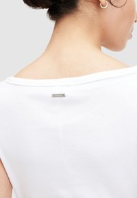 Weiße Baumwollbluse mit schulterfreiem Design. Sie verfügt über ein silbernes Logo-Tag auf der Rückseite und hat eine glatte Textur mit einem einfachen, minimalistischen Look.