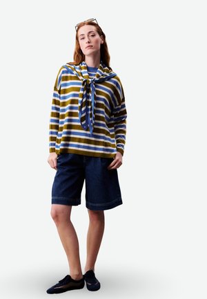 Frau trägt ein gestreiftes Langarmshirt mit passendem Pullover, der über den Schultern gebunden ist, dunkle kniehohe Shorts und schwarze Netzschuhe, steht.