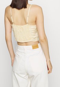 Cropped yellow gingham top with shirred texture, thin straps, worn with white high-waisted jeans featuring a leather label on the back pocket.