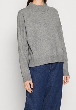 Femme portant un pull en maille gris clair à col montant et manches longues, associé à un jean large bleu foncé.