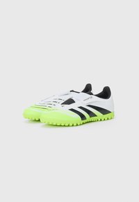Zapatos de fútbol sintéticos blancos con rayas negras; suela de goma verde brillante; recubrimiento texturizado y correa ajustable en la parte frontal.