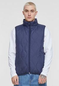 Navy quilted vest with a high collar and full front zipper. Features wave-like stitching and is layered over a white long-sleeve shirt.