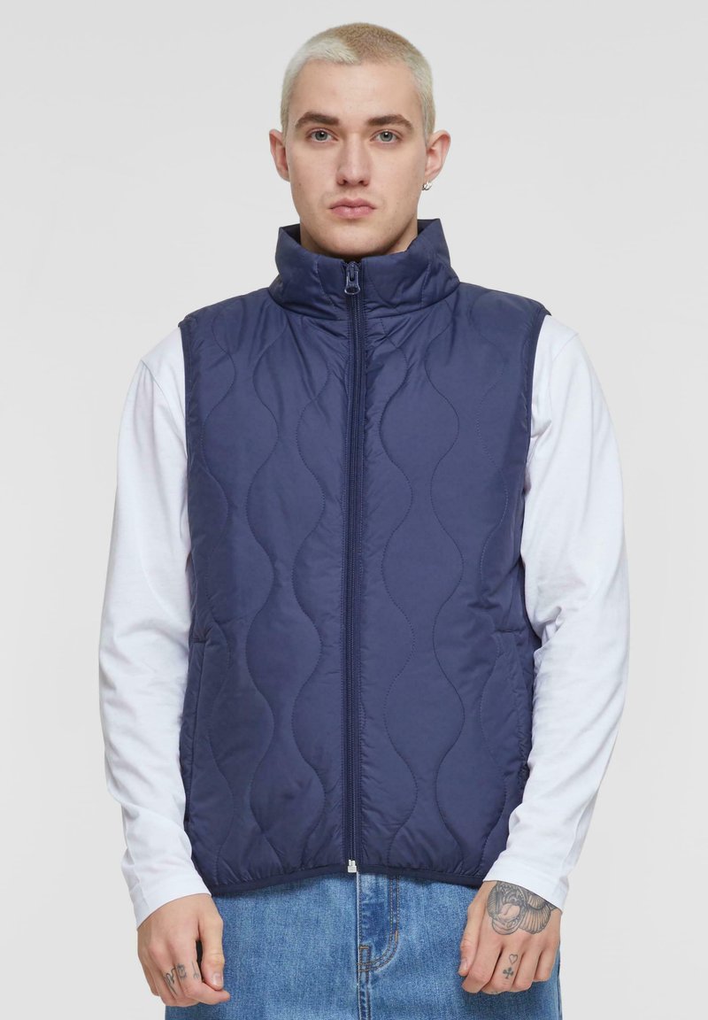 Navy quilted vest with a high collar and full front zipper. Features wave-like stitching and is layered over a white long-sleeve shirt.