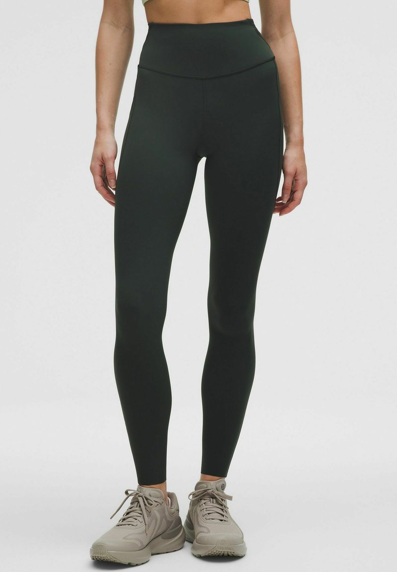 Legginsy w kolorze ciemnozielonym, o wysokim stanie, w bezszwowym wzornictwie, wykonane z elastycznego materiału; zestawione z jasnoszarymi butami sportowymi.