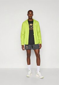 Lichtgewicht, felgroene jas met ritssluiting, gedragen over een zwart shirt met berglogo, gecombineerd met zwart kort broekje met patroon en witte sneakers.