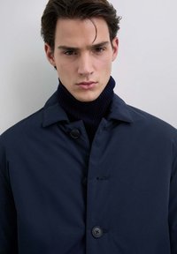 Joven con cabello oscuro que lleva un abrigo azul marino y un suéter de cuello alto negro, mirando al frente contra un fondo claro y liso.