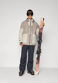 Mees suusajakis, pükstes, saapastes ja prillides hoiab püsti suusapaari lihtsa tausta ees.