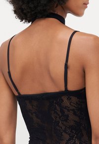 O bodysuit de renda preta apresenta um design de pescoço halter, tiras finas ajustáveis e um padrão texturizado com motivos florais nas costas.