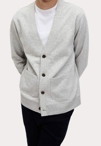 Cardigan grigio chiaro realizzato in tessuto morbido, con scollo a V, chiusura con bottoni e due tasche anteriori. Decorato con bottoni marroni scuri.