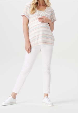 Femme enceinte portant un haut rayé à manches courtes et un pantalon blanc, debout avec une main sur le ventre et les jambes croisées, sur un fond uni.