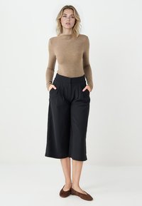 Jasnobrązowa bluzka z długim rękawem zestawiona z czarnymi spodniami o szerokich nogawkach, które mają kieszenie. Stylizacja jest prosta, z gładkimi fakturami i czystymi liniami.