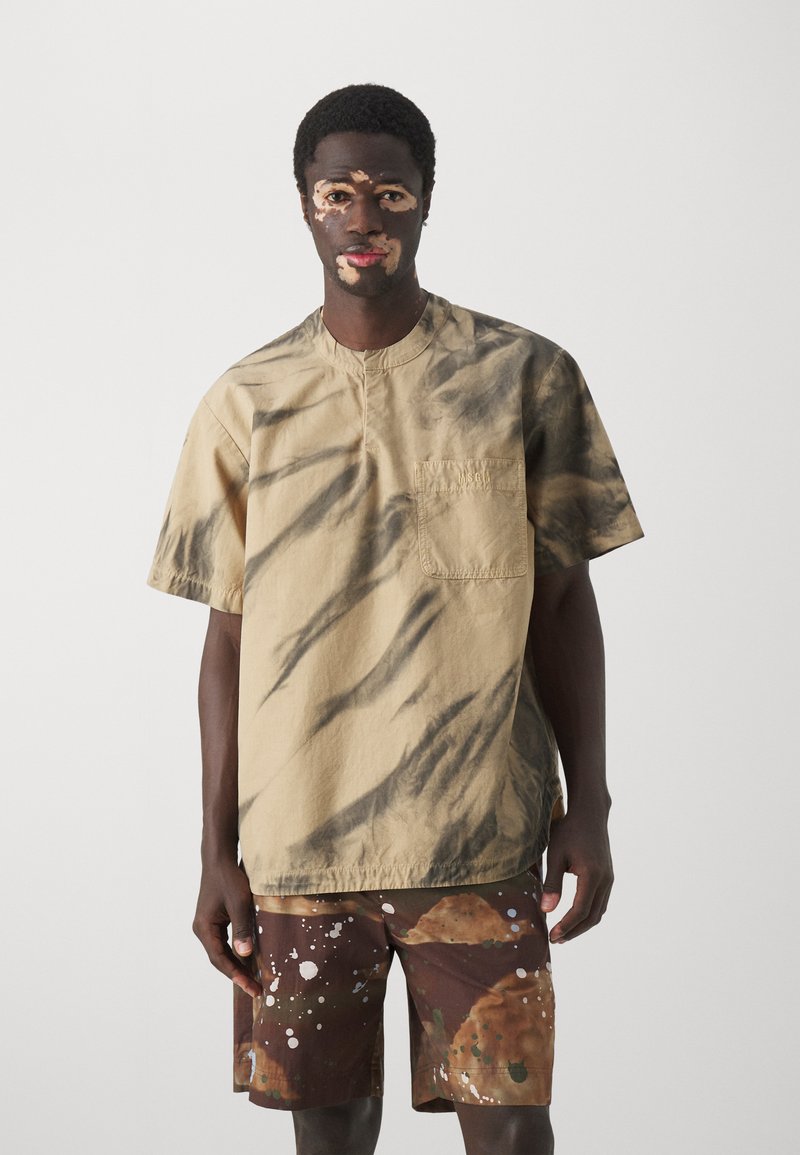Kortærmet, beige skjorte med sorte tie-dye mønstre, med en brystlomme. Sammen med brune shorts, der har farverige splatter-designs.