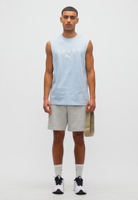 Άντρας που στέκεται, φοράει ανοιχτό μπλε αμάνικο πουκάμισο με το κείμενο "HYROX", γκρι σορτς, λευκές κάλτσες και λευκά αθλητικά παπούτσια, κρατώντας μια μπεζ λάστιχο αντίστασης.