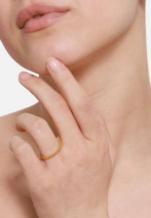 Anello d'oro con piccole palline sferiche, indossato sull'indice. La pelle ha un tono morbido e le unghie sono naturali, con uno sfondo uniforme.