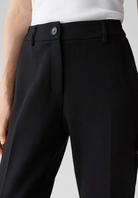 Gros plan sur un pantalon noir sur mesure avec une taille boutonnée, porté avec un haut blanc côtelé.