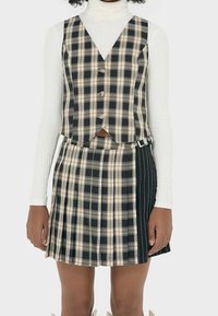 Ensemble de gilet et jupe en tartan avec un mélange de motifs noirs, beiges et blancs. Le gilet a une coupe courte, et la jupe présente des plis et une boucle sur le côté.