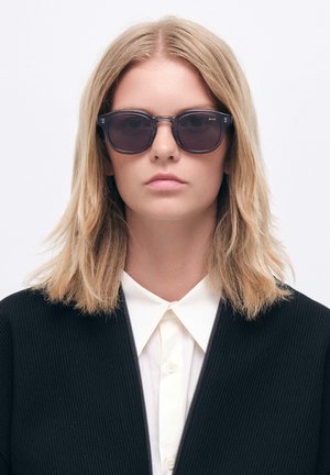 Sorte ovale solbriller med mørke linser, der har en gennemsigtig ramme. Modellen har en sort ribbede jakke over en hvid skjorte med krave.