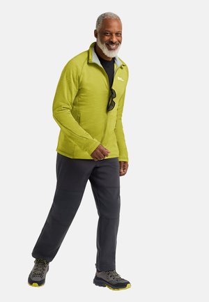 Grüne Fleecejacke mit einem hohen Kragen und Reißverschluss, kombiniert mit grauen Hosen und robusten schwarzen Schuhen mit gelben Akzenten und Schnürsenkeln.