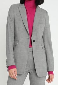 Šedý blazer s přizpůsobeným střihem, s klopovými límci a dvěma předními kapsami. Nosený přes růžový rolák, dodává kontrast a texturu.