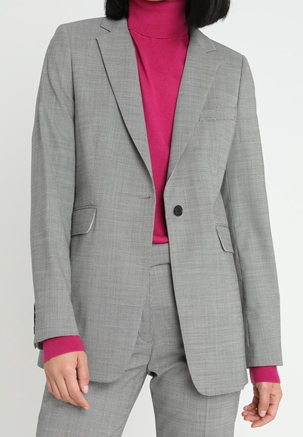 Šedý blazer s přizpůsobeným střihem, s klopovými límci a dvěma předními kapsami. Nosený přes růžový rolák, dodává kontrast a texturu.