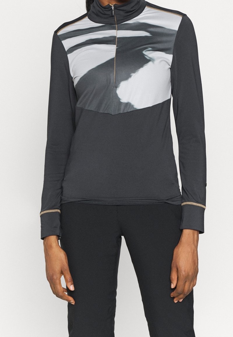 Personne portant un pull noir à manches longues avec une fermeture éclair à moitié ouverte et un motif abstrait en noir et blanc traversant la poitrine et les épaules.