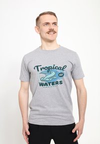 Camiseta de algodón gris con un diseño gráfico que presenta el texto "AGUAS TROPICALES" y una ilustración de un tiburón martillo. Mangas cortas.