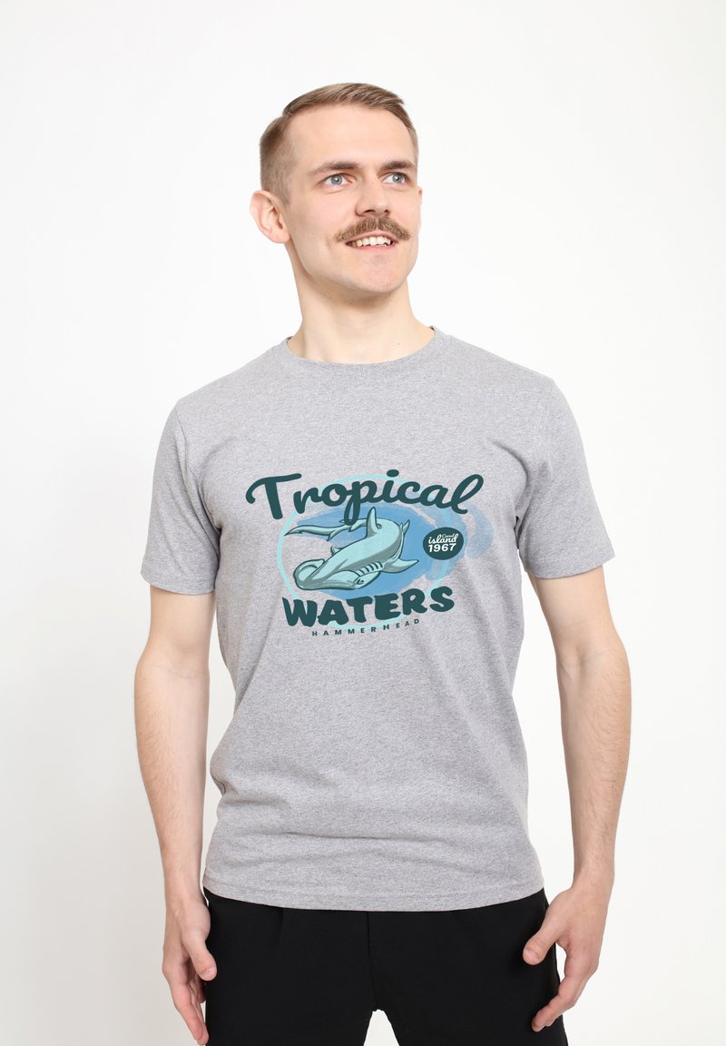 Camiseta de algodón gris con un diseño gráfico que presenta el texto "AGUAS TROPICALES" y una ilustración de un tiburón martillo. Mangas cortas.