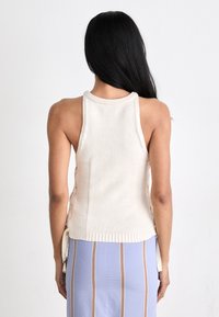 Πλεκτό τοπ με σκίσιμο στον λαιμό σε κρεμ απόχρωση με υφή, λεπτομέρειες κορδονιού στο πλάι και ριμπ τελείωμα. Συνδυάζεται με φούστα με λευκές και μωβ ρίγες.