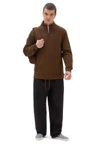 Felpa marrone con zip e colletto piatto, tasca a marsupio e orlo con cordino, abbinata a pantaloni neri e scarpe slip-on a quadretti.