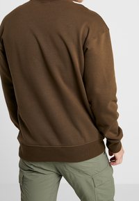 Homme portant un sweat-shirt à manches longues marron et un pantalon cargo vert clair, vu de dos sur un fond blanc.