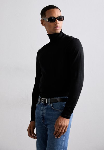 Svart turtleneck-tröja, åtsittande design, bärs med blå jeans och ett svart bälte. Fyrkantiga, mörka solglasögon fulländar looken.