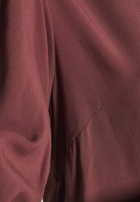 Bordowy materiał o miękkiej fakturze, z drapowanym wzorem i subtelnymi plisami wzdłuż szwu, przedstawiony na zdjęciu zbliżeniowym.