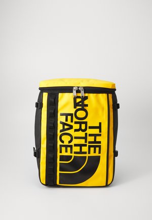 Gelbes und schwarzes Rucksack mit Reißverschluss, ausgestattet mit großem Logo-Print, verstellbaren Seitenträgern und einem zylindrischen Design. Glatte Textur.