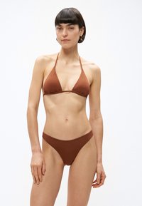 Γυναίκα με κοντά σκουρόχρωμα μαλλιά που φοράει καφέ τρίγωνο μπικίνι, στέκεται μπροστά σε λευκό απλό φόντο.