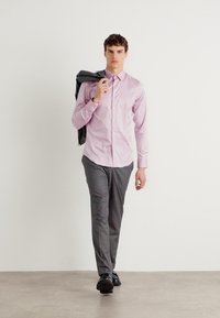 Helles rosa Hemd mit Knopfleiste, graue Hose und schwarze Schuhe. Das Model trägt einen dunklen Blazer und geht vor einem einfarbigen Hintergrund.