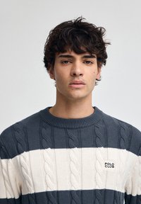 Sweter z dzianiny w ciemnoszarym i kremowym kolorze, z poziomymi paskami, teksturą dzianiny w warkocze oraz małą metką 'POLO CLUB' na piersi.