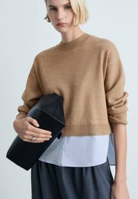 Brauner Strickpullover mit geripptem Saum, über einem weißen Hemd getragen, kombiniert mit einer schwarzen Clutch. Weiche Textur, lässiger Schnitt und strukturierte Form.
