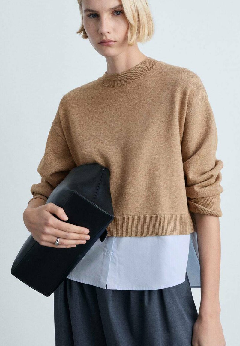 Brauner Strickpullover mit geripptem Saum, über einem weißen Hemd getragen, kombiniert mit einer schwarzen Clutch. Weiche Textur, lässiger Schnitt und strukturierte Form.