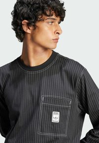 Black long-sleeve shirt featuring vertical white pinstripes, round neckline, and a front pocket with a white brand label. Smooth texture.