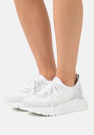 Baskets blanches à lacets avec semelle texturée portées pieds nus, vues de profil sur un fond clair uni.