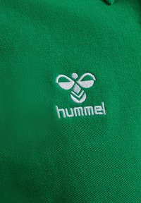 Grön polotröja i texturerat material med en vit broderad logotyp av en bi och ordet "hummel" under.