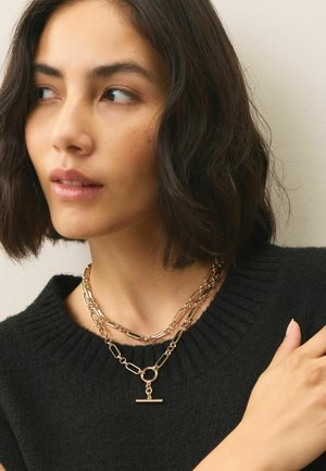 Collane a catena a più livelli in tono oro con diverse forme di anelli. Include un gancio circolare e un accento a barra orizzontale. Collo di un maglione nero visibile.