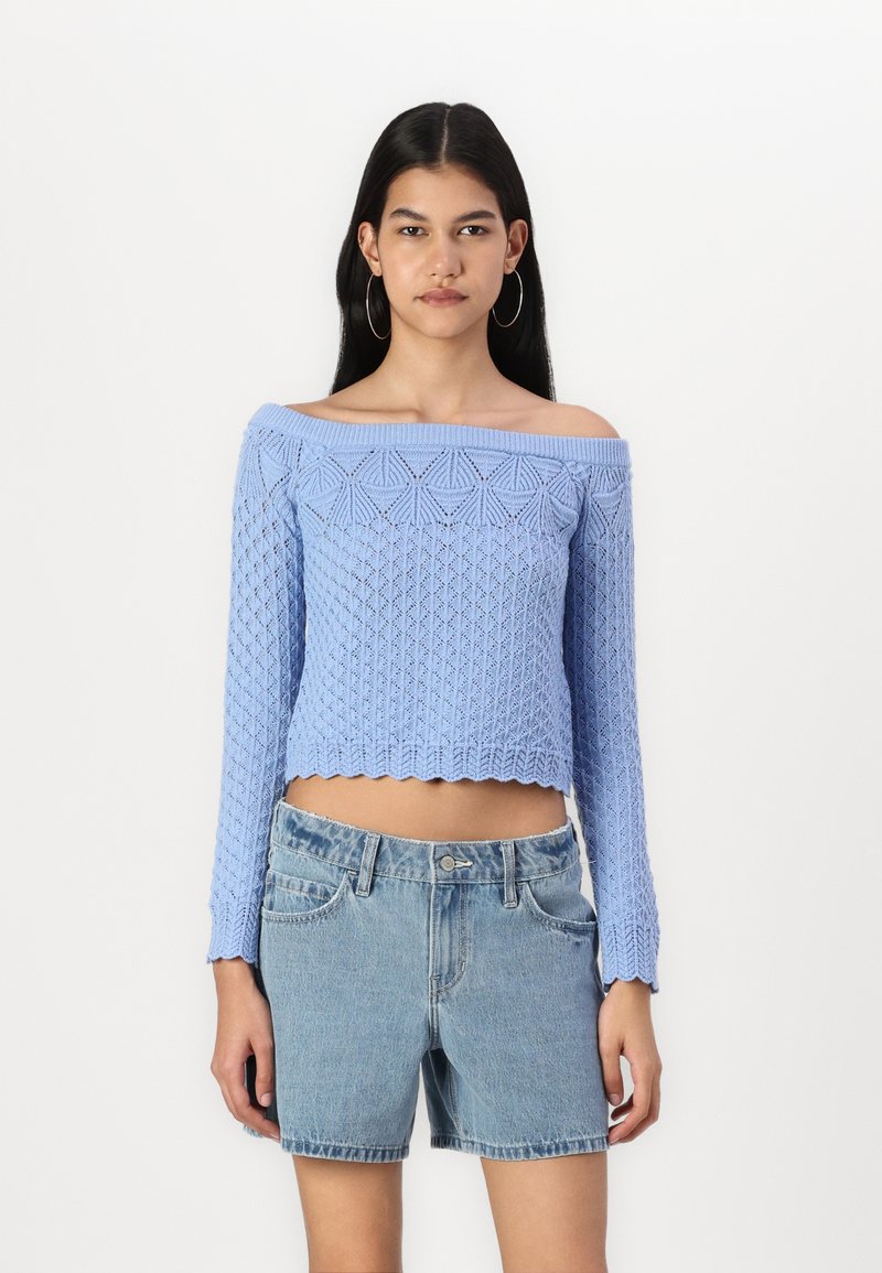 Ljusblå, off-the-shoulder stickad tröja med strukturerade mönster; kombinerad med högmidjade denimshorts. Vågig fåll och långa ärmar.