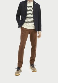 Blazer azul marino sobre un suéter de rayas color crema, pantalones marrones y zapatillas beige con cordones azules. Se aprecian tejidos texturizados y un ajuste a medida.