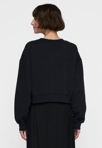 Sudadera corta negra con mangas largas holgadas, cuello y puños acanalados, y textura suave, vista desde atrás.