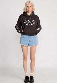 Zwarte capuchontrui met witte print, spijkershorts en zwarte schoenen. Model staat met handen in de zakken en toont een casual outfit.