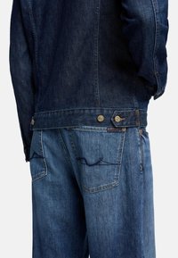 Giacca in denim scuro con chiusure a bottoni, abbinata a jeans in denim chiaro con dettagli di cucitura nelle tasche e una vestibilità comoda.