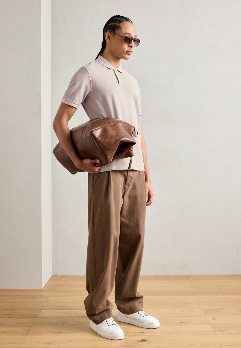 Beżowa koszulka polo, luźne brązowe spodnie i białe trampki. Trzyma dużą, ciemno-brązową skórzaną torbę o eleganckim designie i metalowymi akcentami.